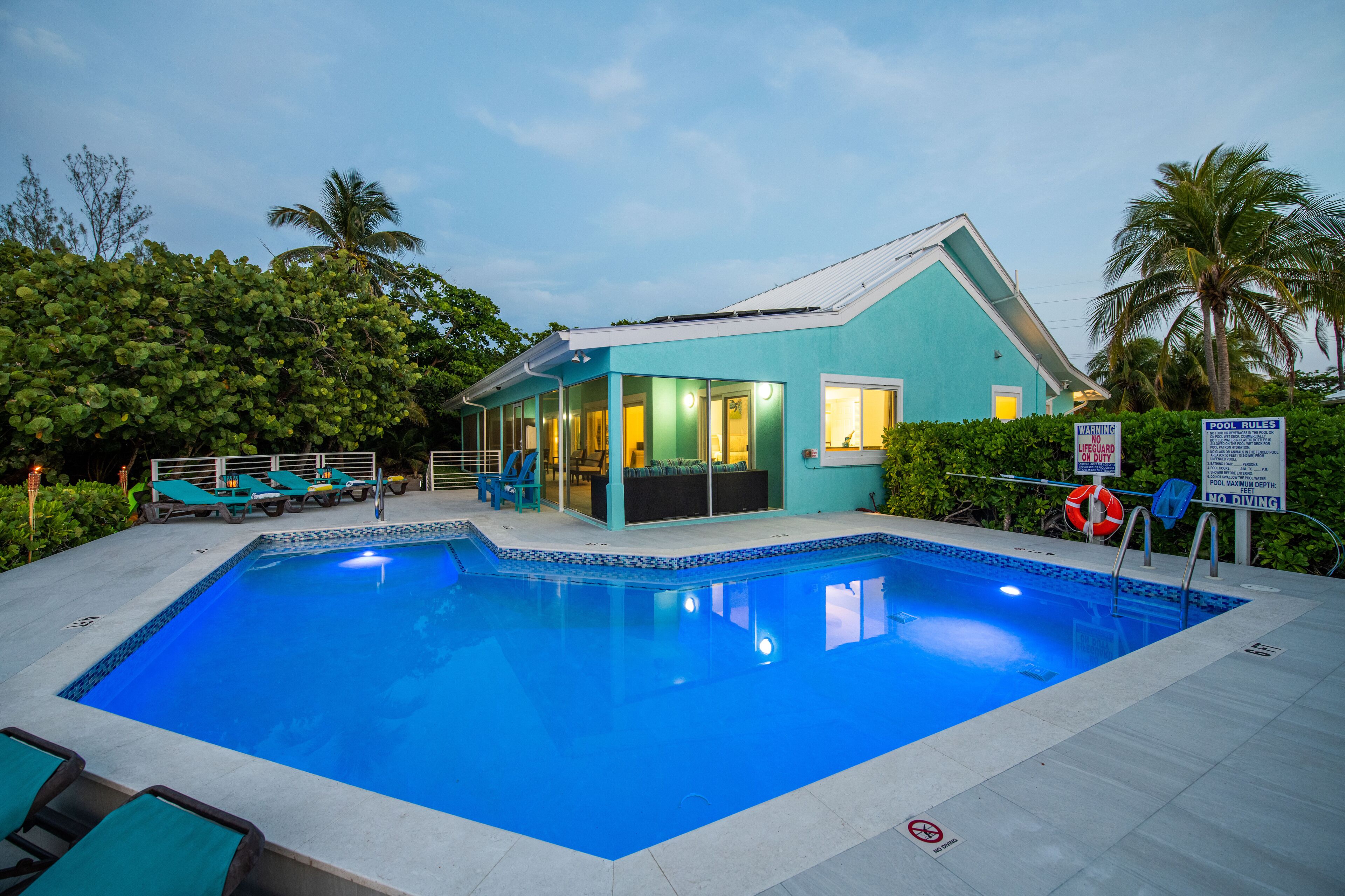 Una piscina con lettini, una casa con facciata blu e palme sullo sfondo.