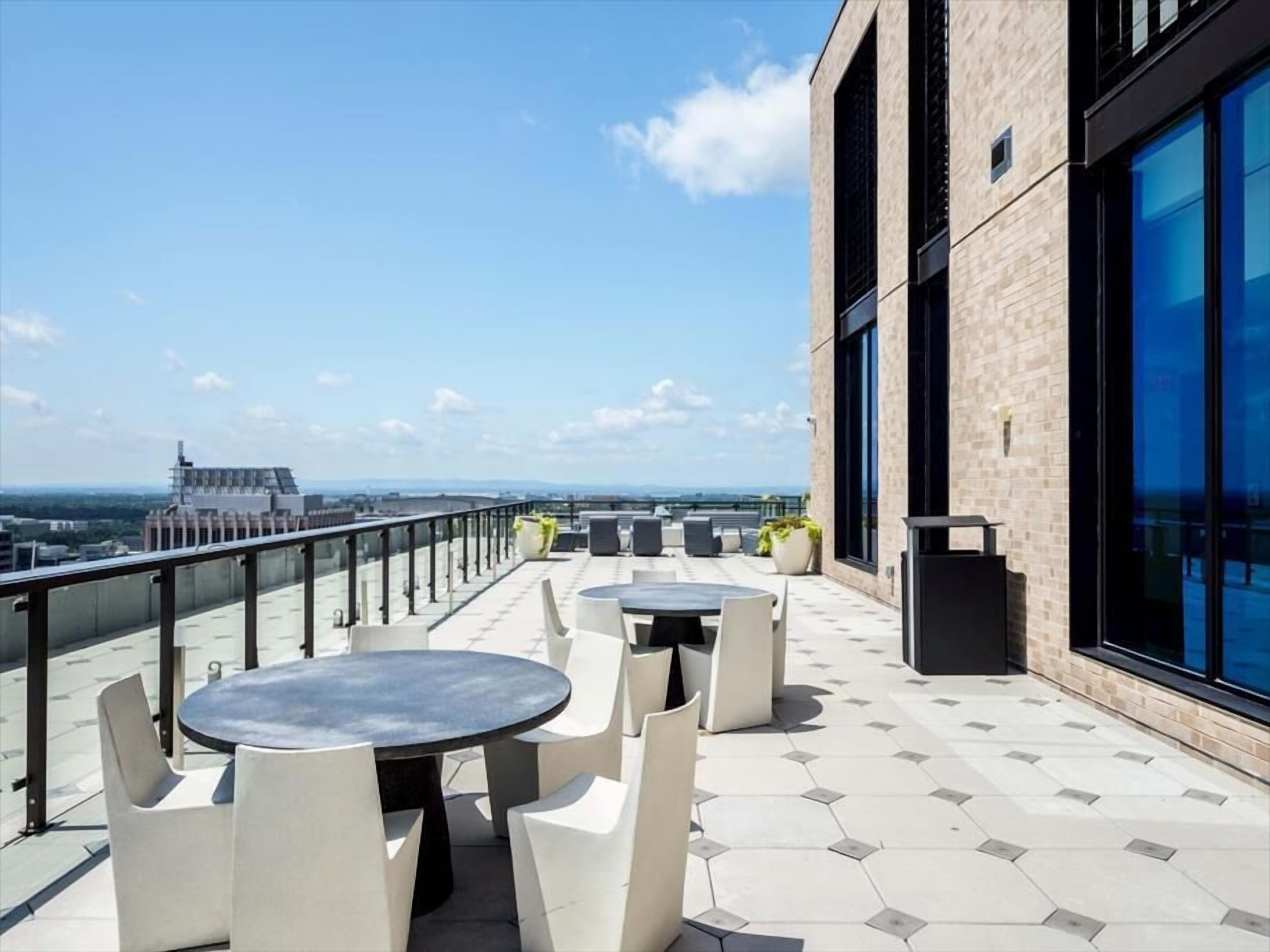 A rooftop terrace with round tables and chairs, a glass balustrade, and a view of the cityscape.