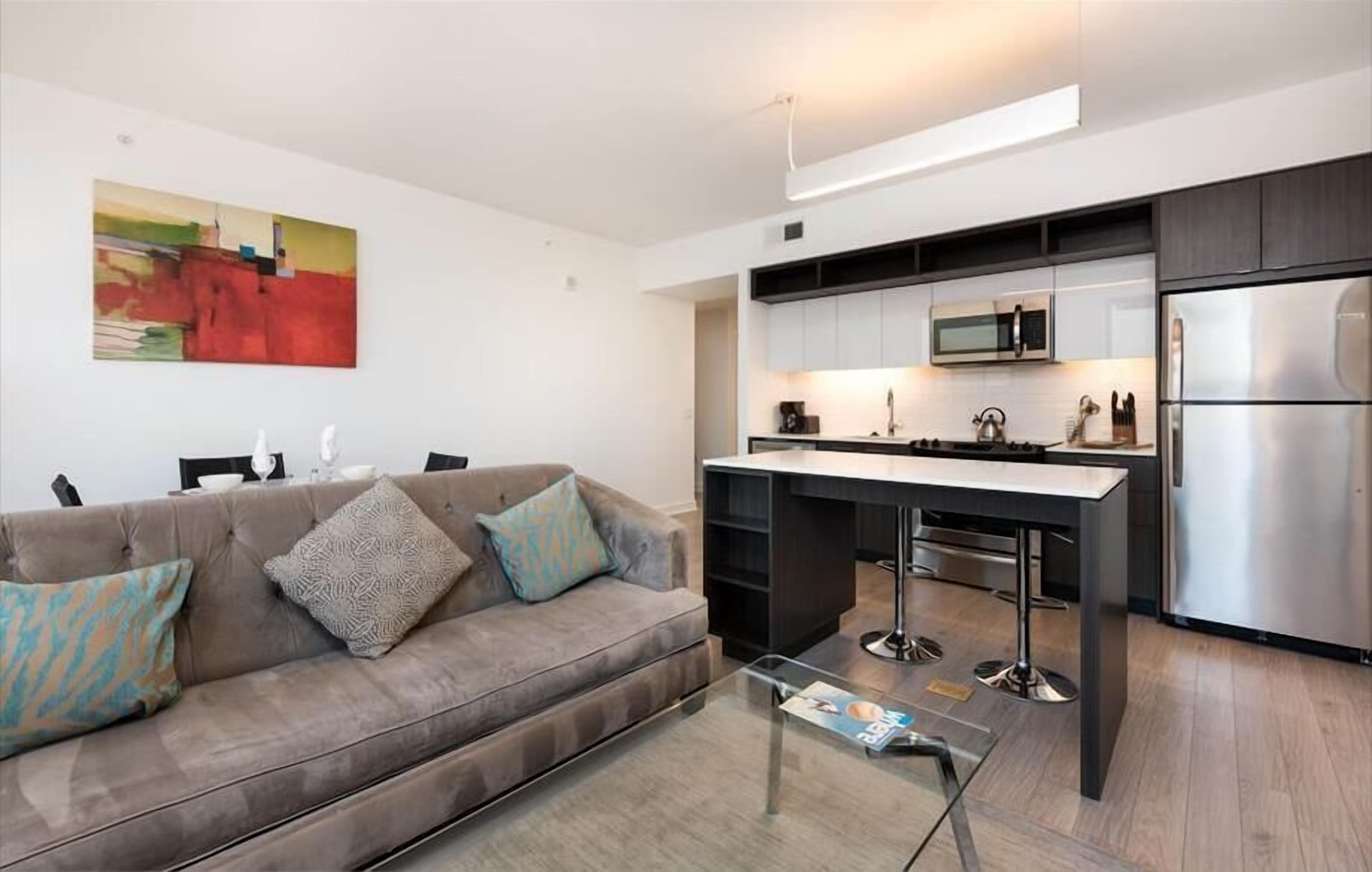 A modern apartment with a kitchenette, a sofa with patterned cushions, and a glass coffee table.