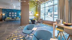 A modern hotel lobby with a blue and yellow patterned carpet, a seating area with blue chairs, a dining table with a metal bucket and fruit, and a large window offering a city view. - Near NIH | Full Kitchen | Central Farm Market Nearby | by GLS (Bethesda)