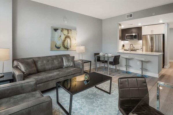 Ein modernes Wohnzimmer mit grauem Sofa, Couchtisch und Essbereich, im Hintergrund eine Küche.