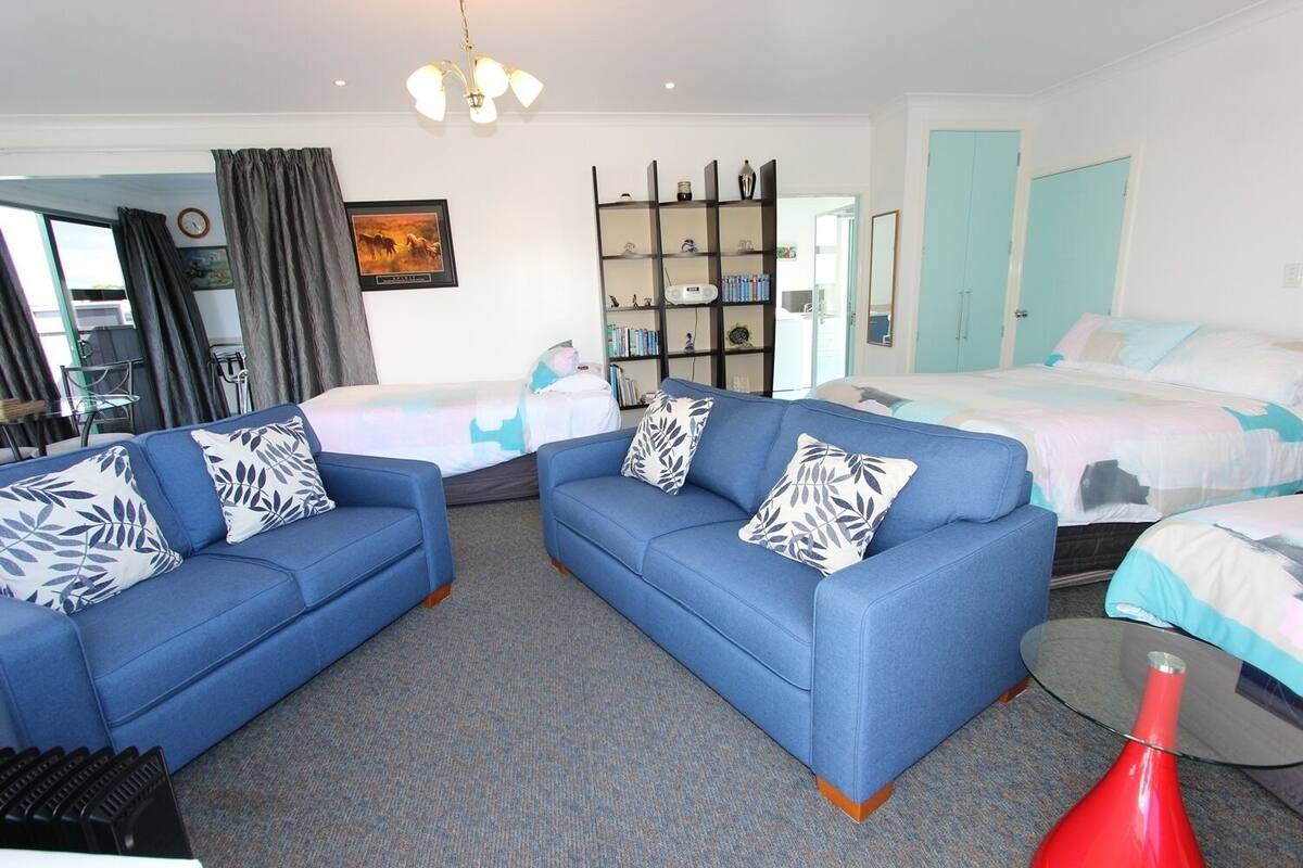 Habitación de hotel con sofá azul, cama con ropa de cama estampada, estantería y un jarrón rojo.