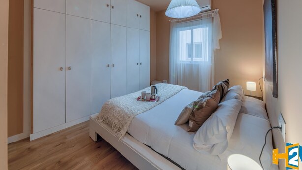 Un dormitorio con un armario grande, una cama con almohadas, una mesita de noche con lámpara y una ventana con cortinas translúcidas.