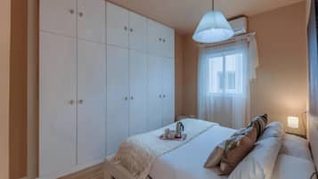 Una camera da letto con un armadio grande, un letto con cuscini, un comodino con una lampada e una finestra con tende leggere.