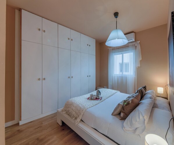 A bedroom with a large wardrobe, a bed with pillows, a bedside table with a lamp, and a window with sheer curtains.