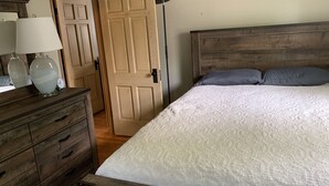 Una camera da letto con un letto in legno, una coperta trapuntata bianca, due cuscini grigi, un comò in legno con uno specchio e una lampada.