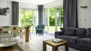 Una sala de estar moderna con un sofá, una mesa de comedor con sillas, una mesa de centro y una silla. Amplias ventanas con cortinas, una lámpara de techo y un aplique de pared.