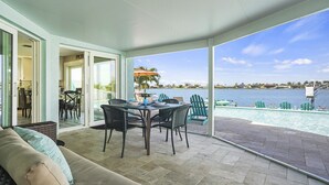 Un patio couvert avec une table et des chaises, un canapé, et une vue sur une piscine et un plan d’eau.