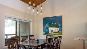 A dining area with a wooden table and chairs, a painting of a house on the wall, and a window with blinds. - SPECTACULAR BALDY VIEW REMODELED 2 BD TOWNHOME NEXT TO LIFTS! LUXURY! LOCATION! (Ketchum)