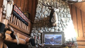 Lareira de pedra com cabeça de veado montada, prateleiras de madeira com troféus de caça e um quadro de uma carruagem puxada a cavalos.
