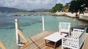 A wooden deck with a white table and two white chairs, overlooking a calm sea with boats and a distant mountain range. - HamO Cosy: Sainte Anne apartment (Sainte Anne)
