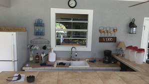 Une cuisine équipée d’un évier, d’un plan de travail et d’une fenêtre. Il y a une horloge au mur et une enseigne sur laquelle on peut lire « Vino ».