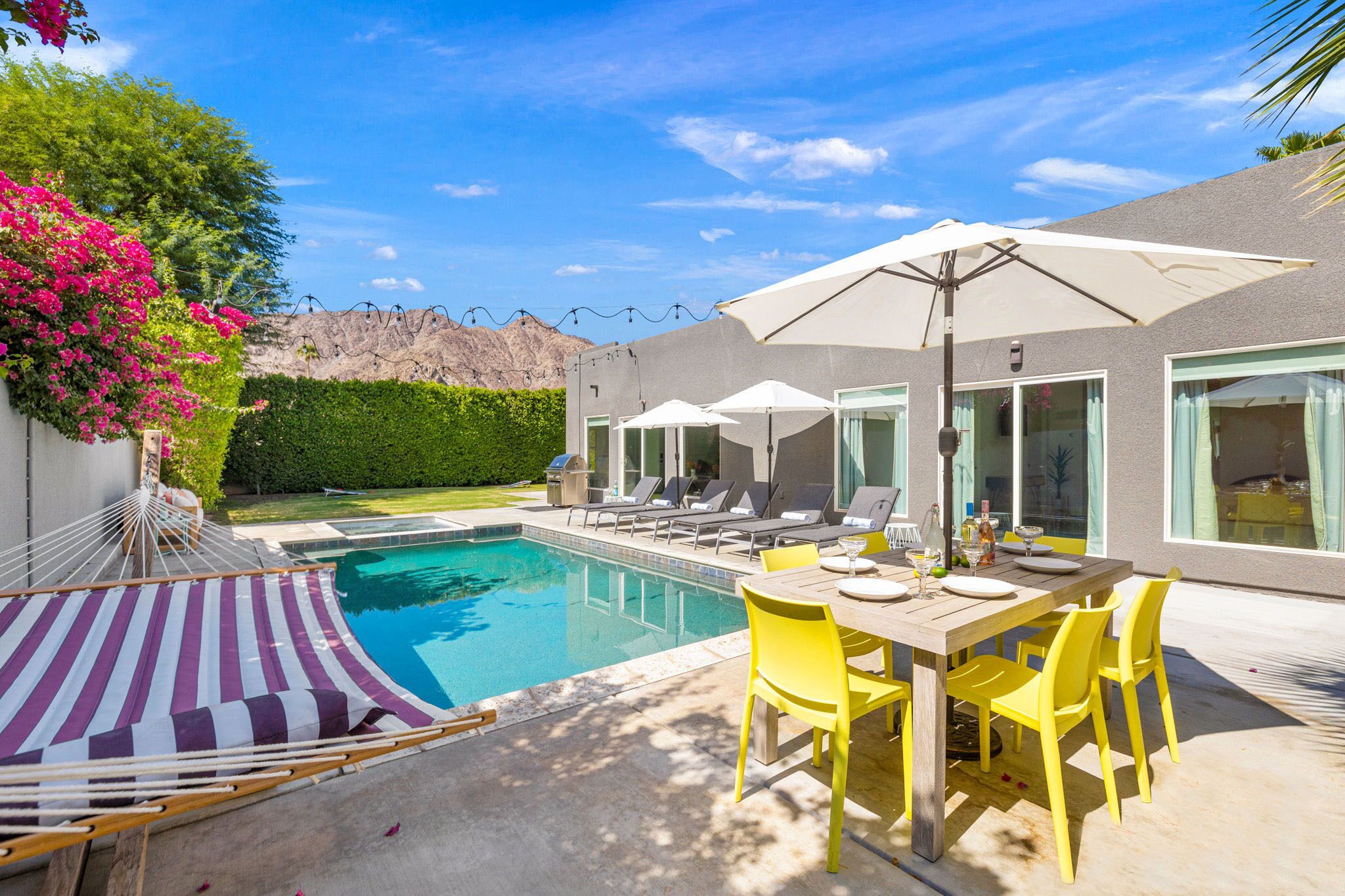 Ein moderner Poolbereich mit Pool, Liegestühlen, einem Esstisch mit gelben Stühlen und einer gestreiften Hängematte.