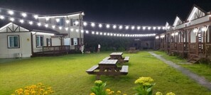 Un patio con mesas de picnic, luces colgantes y un edificio con balcón.
