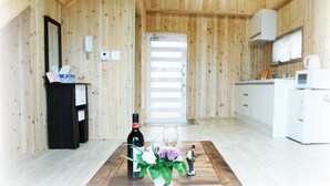 Interior em madeira com uma mesa de jantar, uma garrafa de vinho e um arranjo floral. Há uma área de cozinha com armários e eletrodomésticos brancos.