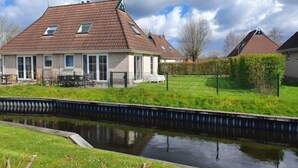 Een huis met een schuin dak, een kleine tuin en een smalle waterloop ervoor.