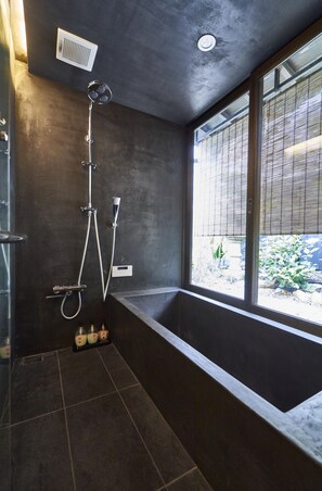 A modern bathroom with a large bathtub, a shower with multiple heads, and a window providing a view of the outdoors. - Zen (Kyoto)