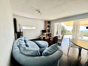 Ein modernes Wohnzimmer mit einer gebogenen Couch, einem Flachbildfernseher, einem Bücherregal und einem Essbereich mit Tisch und Stühlen.