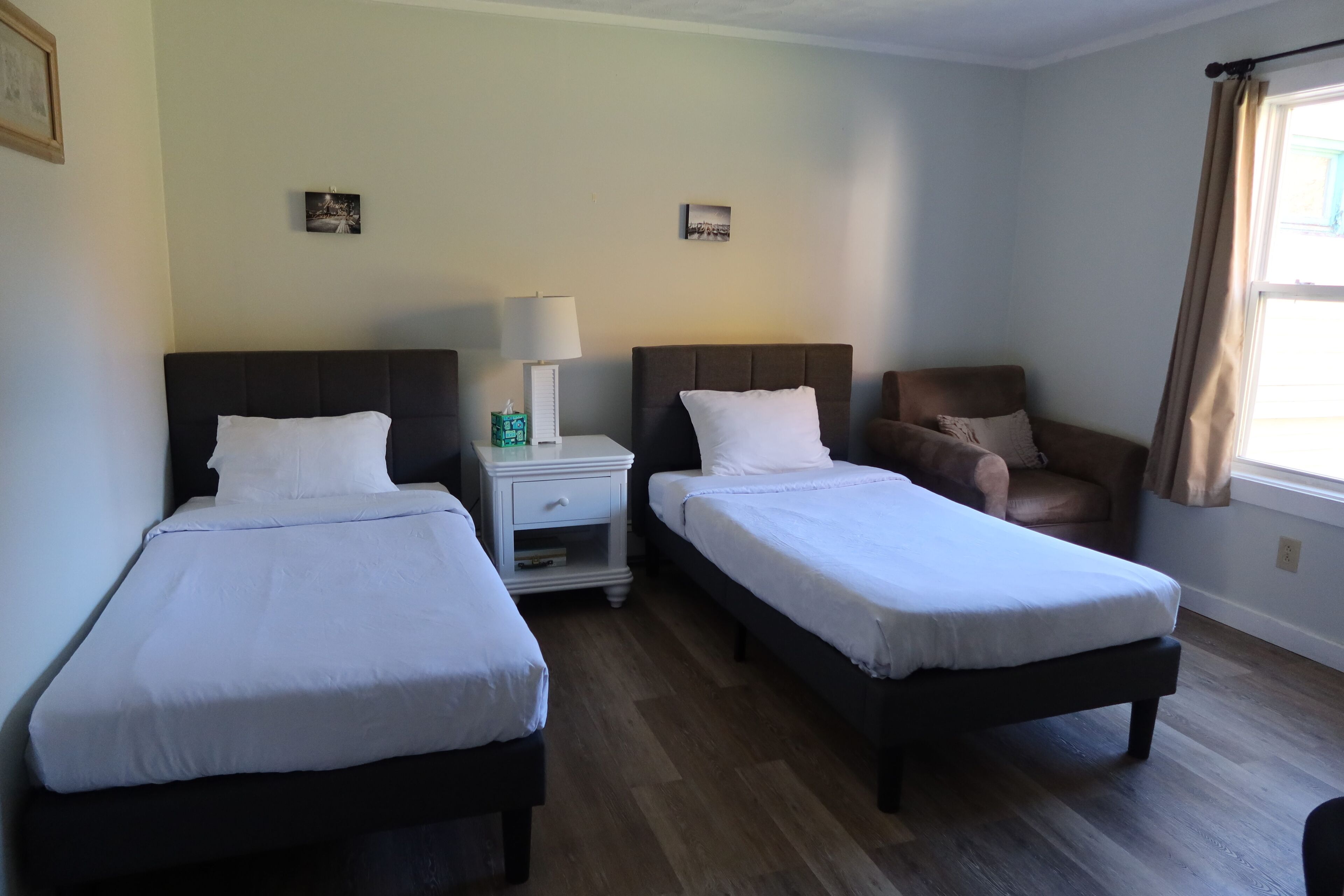 Ein Hotelzimmer mit zwei Einzelbetten, einem Nachttisch mit Lampe, einem Sessel und einem Fenster mit Vorhängen.