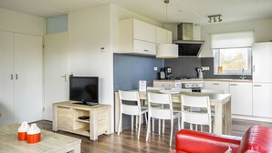 Une cuisine moderne avec des meubles blancs, un coin repas avec des chaises blanches, un canapé en cuir rouge et un téléviseur posé sur une table en bois.