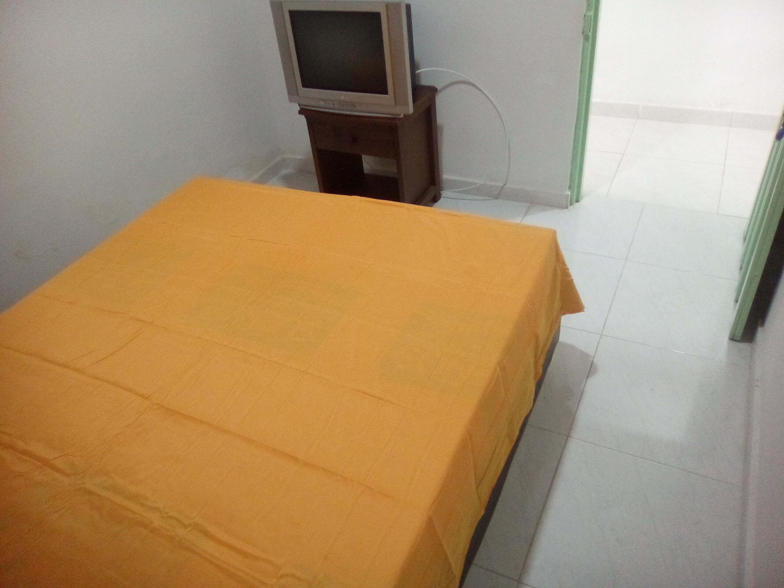 A room with a bed covered in a yellow sheet, a small wooden table with a television on it, and a green door in the background.