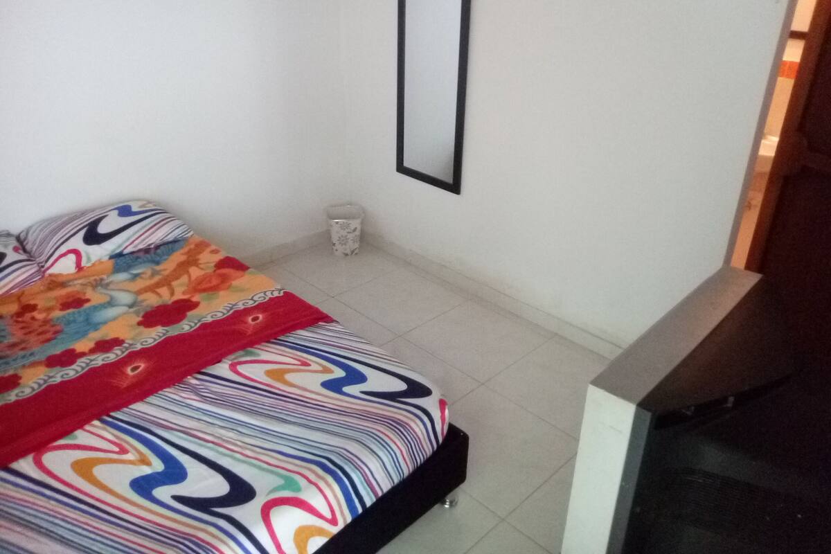 A bedroom with a bed, a bedside table, a mirror, and a trash bin.