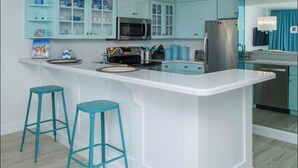Een moderne keuken met turquoise kastjes, een centraal eiland met krukken, roestvrijstalen apparatuur en een kroonluchter.