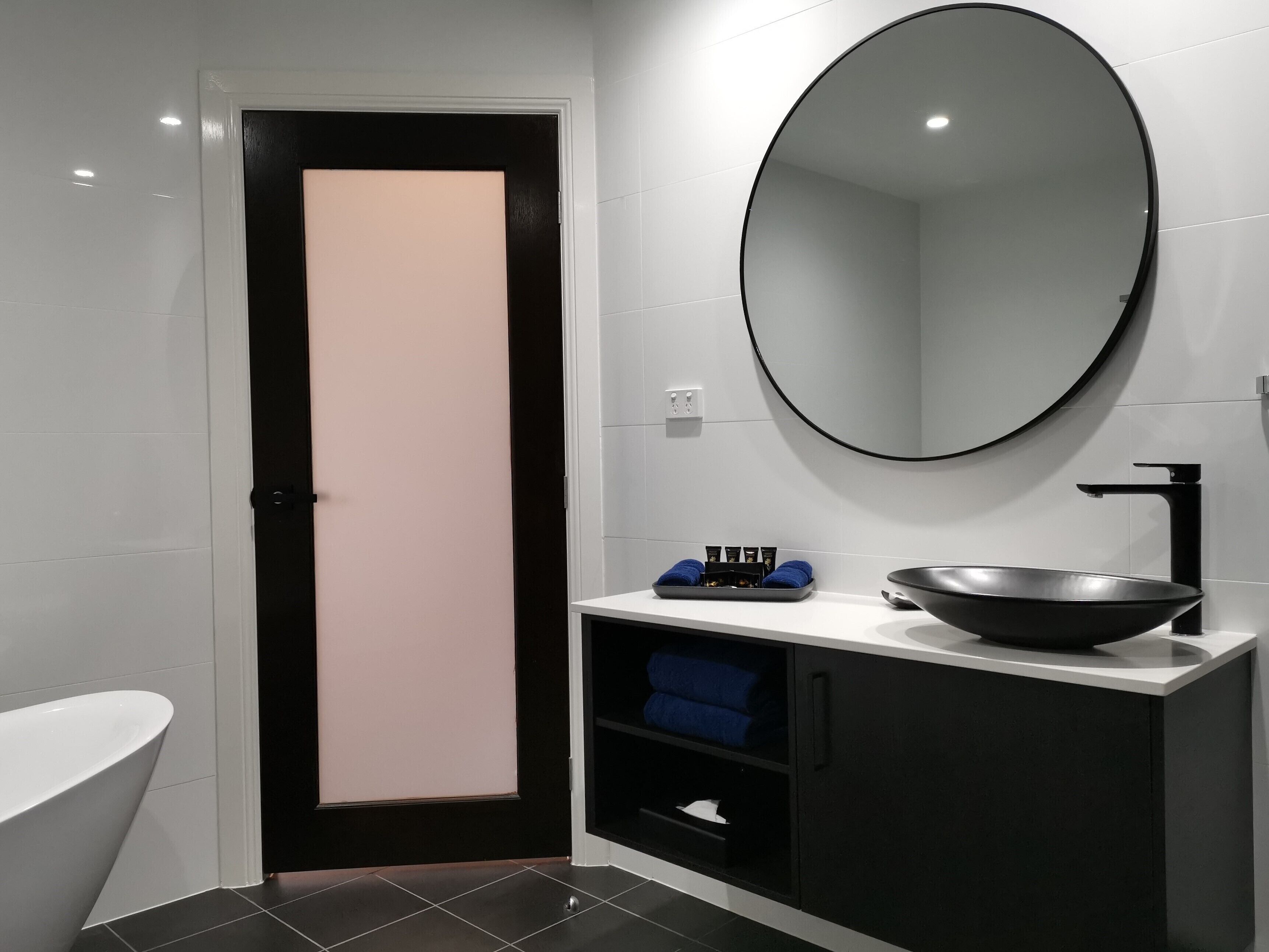 Une salle de bain moderne avec une baignoire sur pieds, un lavabo noir doté d’une grande cuve, un miroir rond et une porte avec un panneau rose.