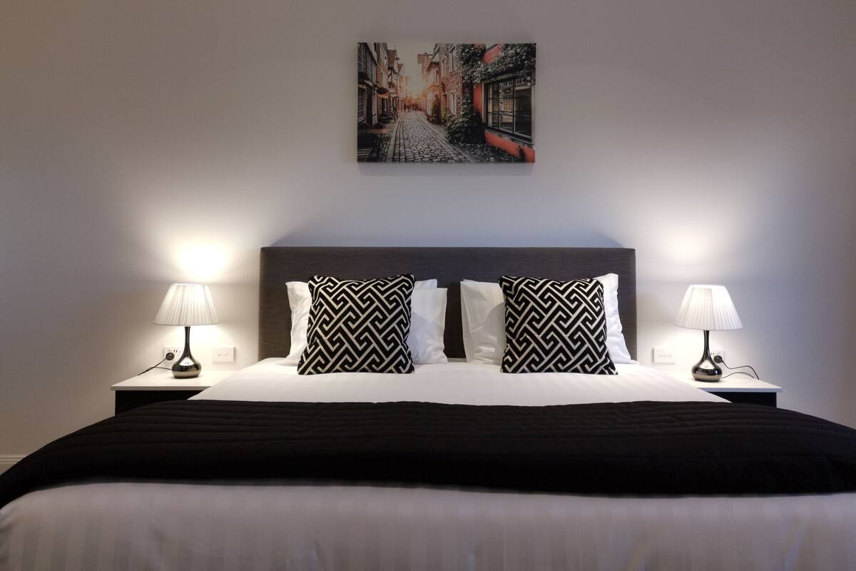 Una cama bien hecha con un cabecero de diseño blanco y negro, dos lámparas de noche y un cuadro enmarcado con una vista urbana sobre la cama.