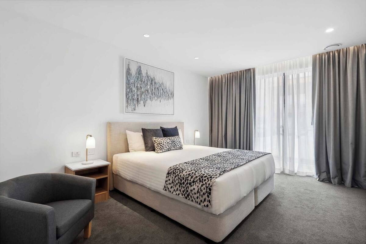 Habitación de hotel moderna con una cama grande, una mesita de noche con lámpara, un sillón gris y un ventanal con cortinas.