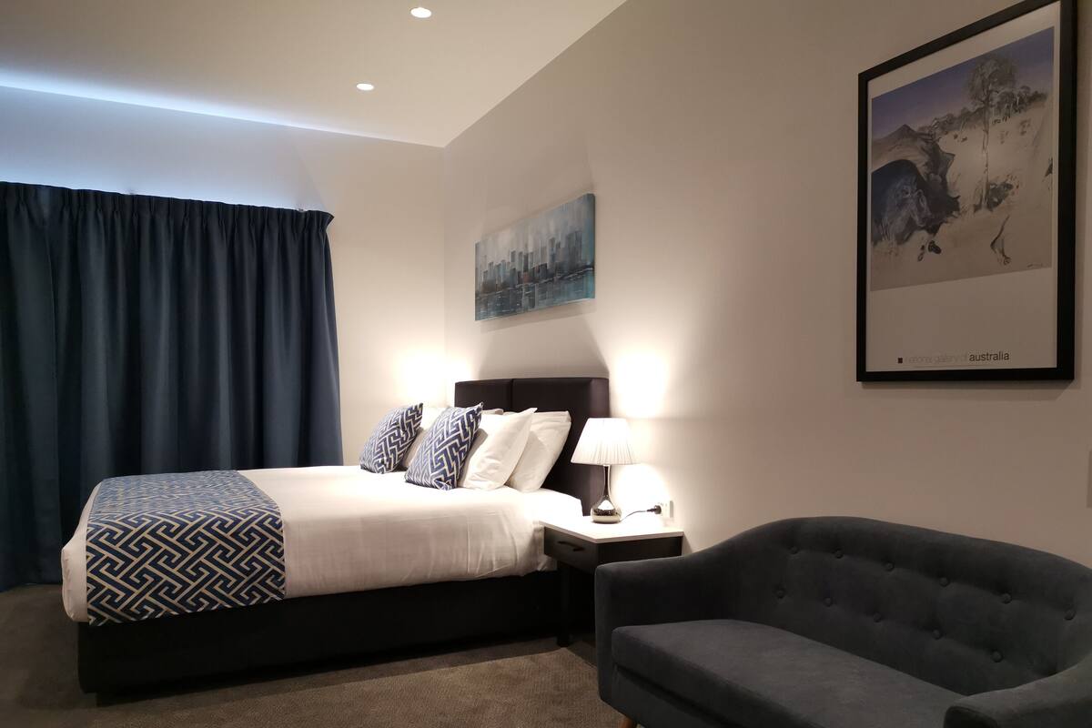 Una habitación de hotel moderna con una cama bien hecha, una mesita de noche con lámpara, un sillón gris y un cuadro en la pared.