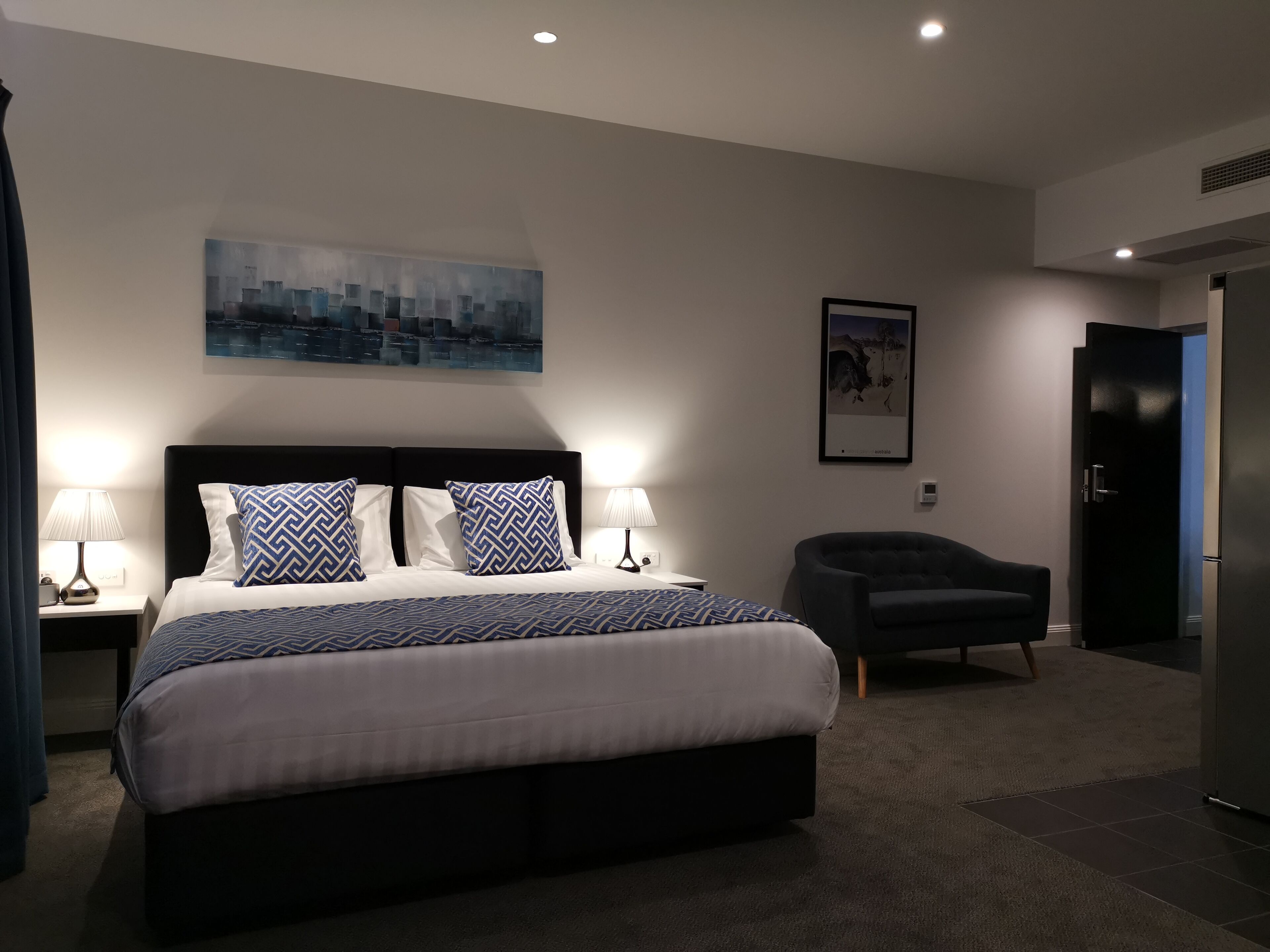 A modern hotel room with a large bed, two bedside lamps, a cityscape painting, a framed picture, a dark armchair, and a closed door.