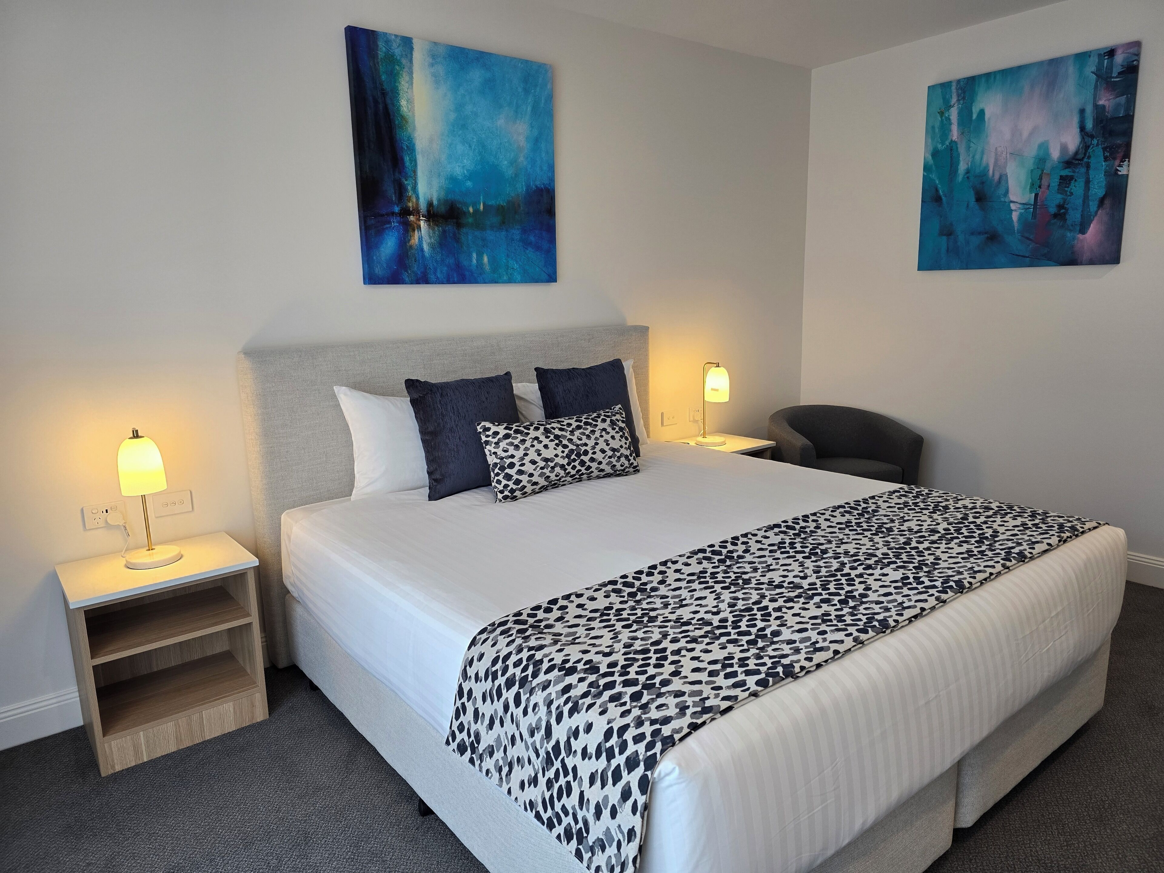 A hotel room with a large bed, two bedside tables, a chair, and two abstract paintings on the walls.