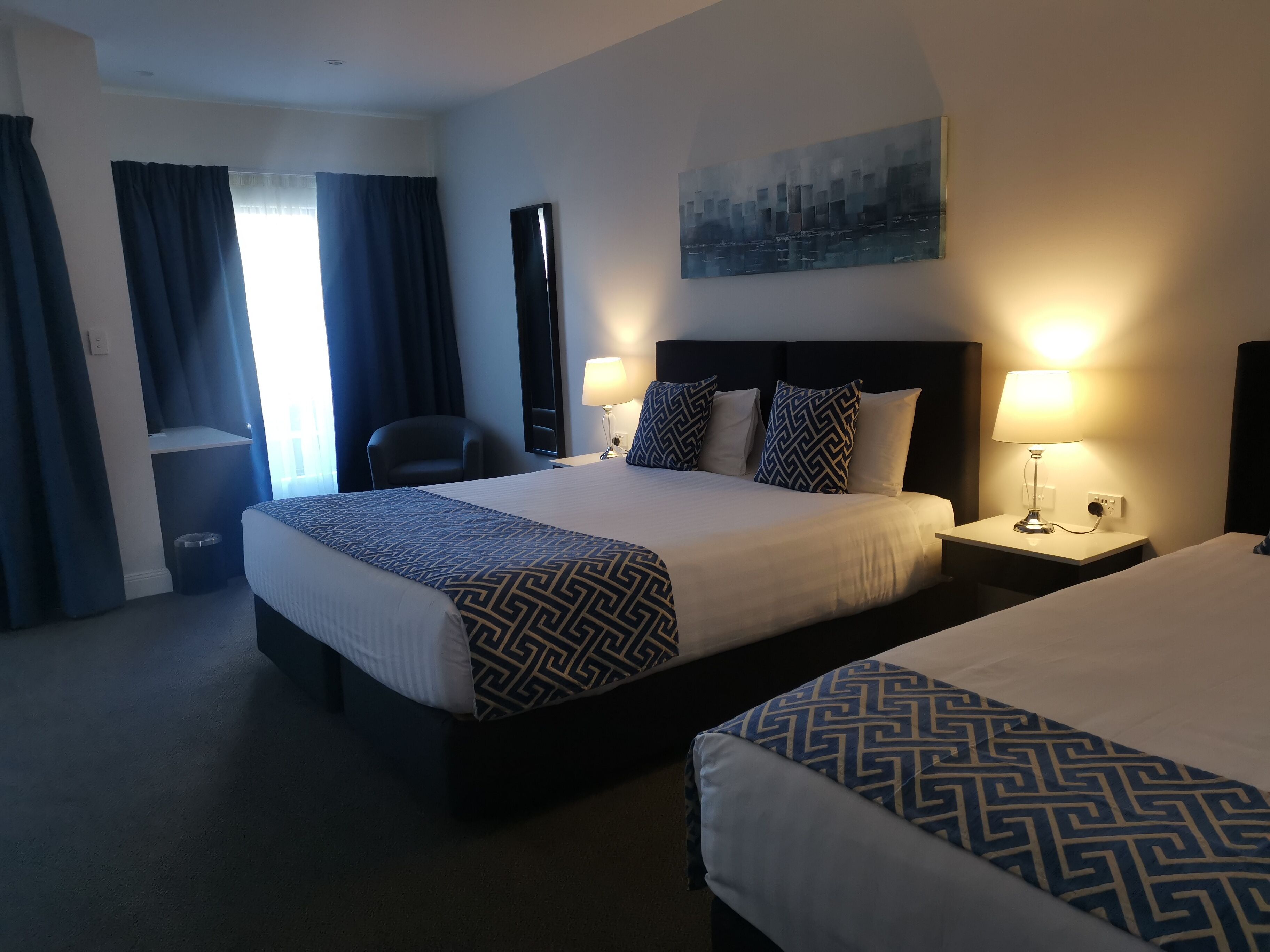 Habitación de hotel con dos camas, un escritorio, una silla, una lámpara y un cuadro en la pared.