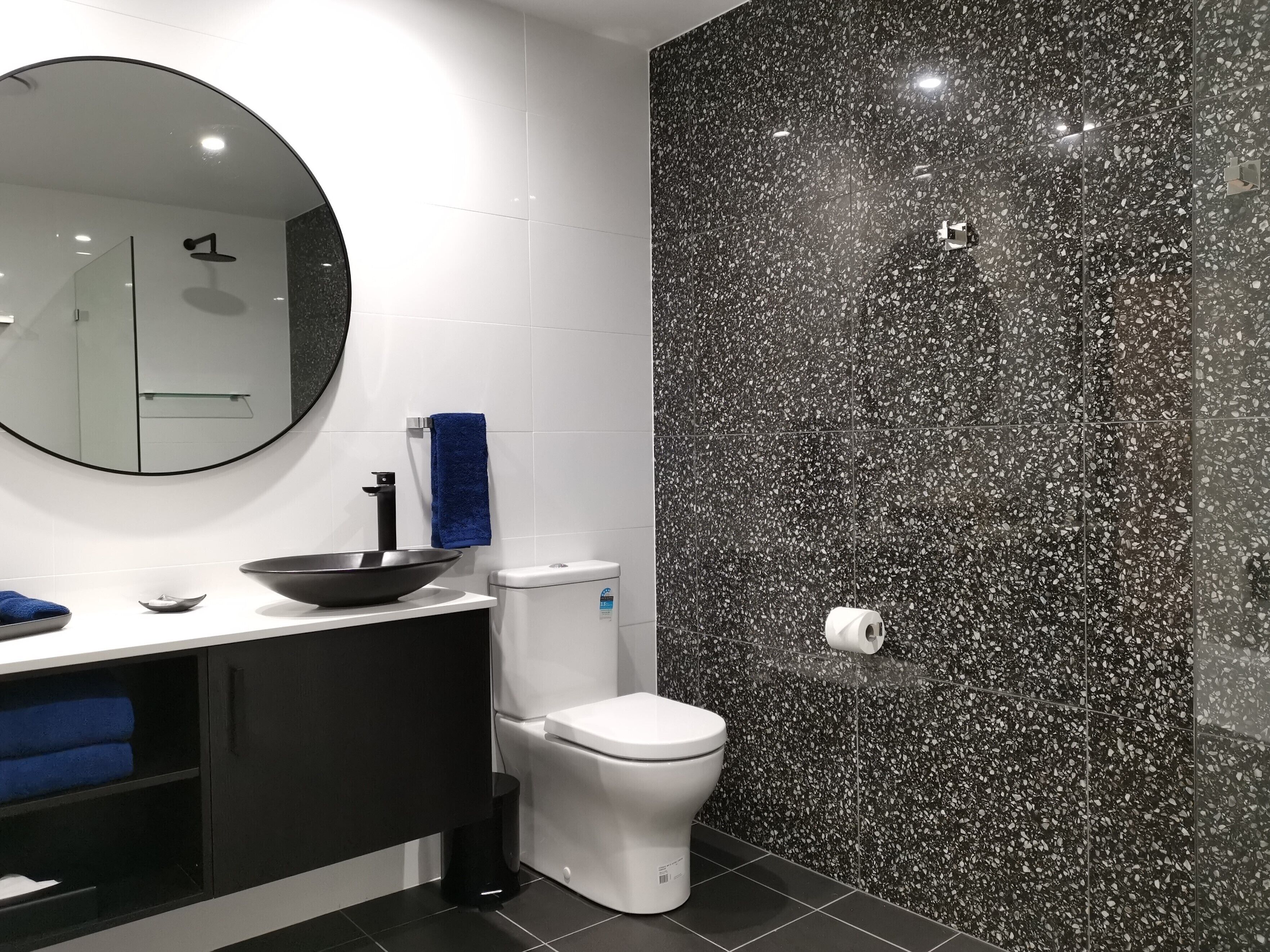 Een moderne badkamer met een wand betegeld met zwart gemêleerd mozaïek, een wit toilet, een zwarte wasbak met kom, een blauwe handdoek en een ronde spiegel.