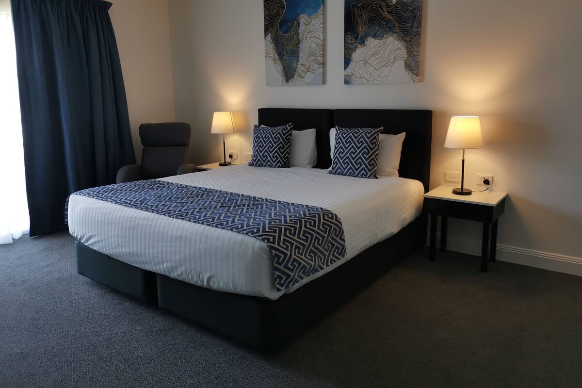 Habitación de hotel con una cama grande, dos lámparas de noche, una silla y dos cuadros abstractos en la pared.