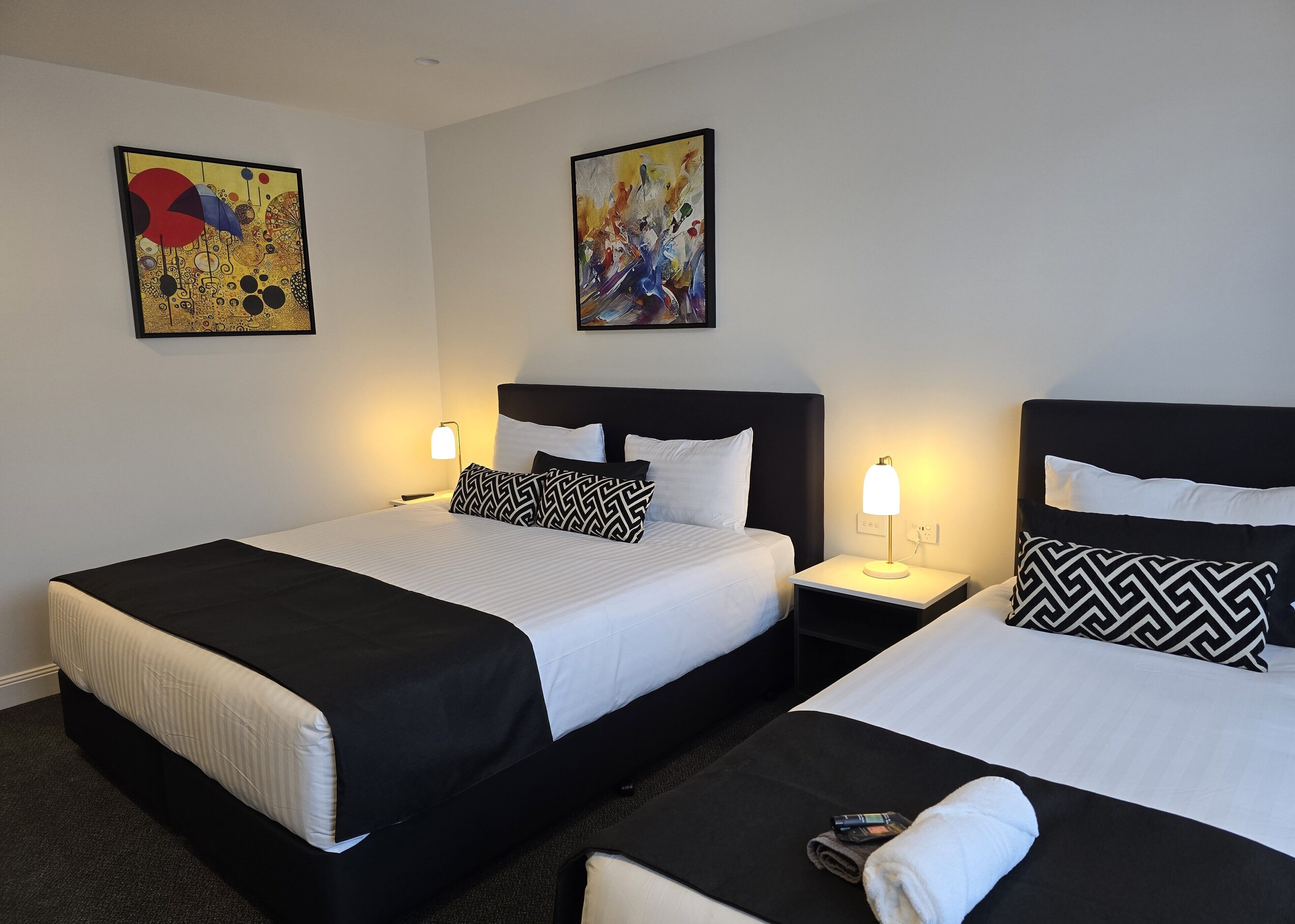 A hotel room with two beds, each with black and white bedding, a black headboard, and a bedside table with a lamp. There are two abstract paintings on the walls.