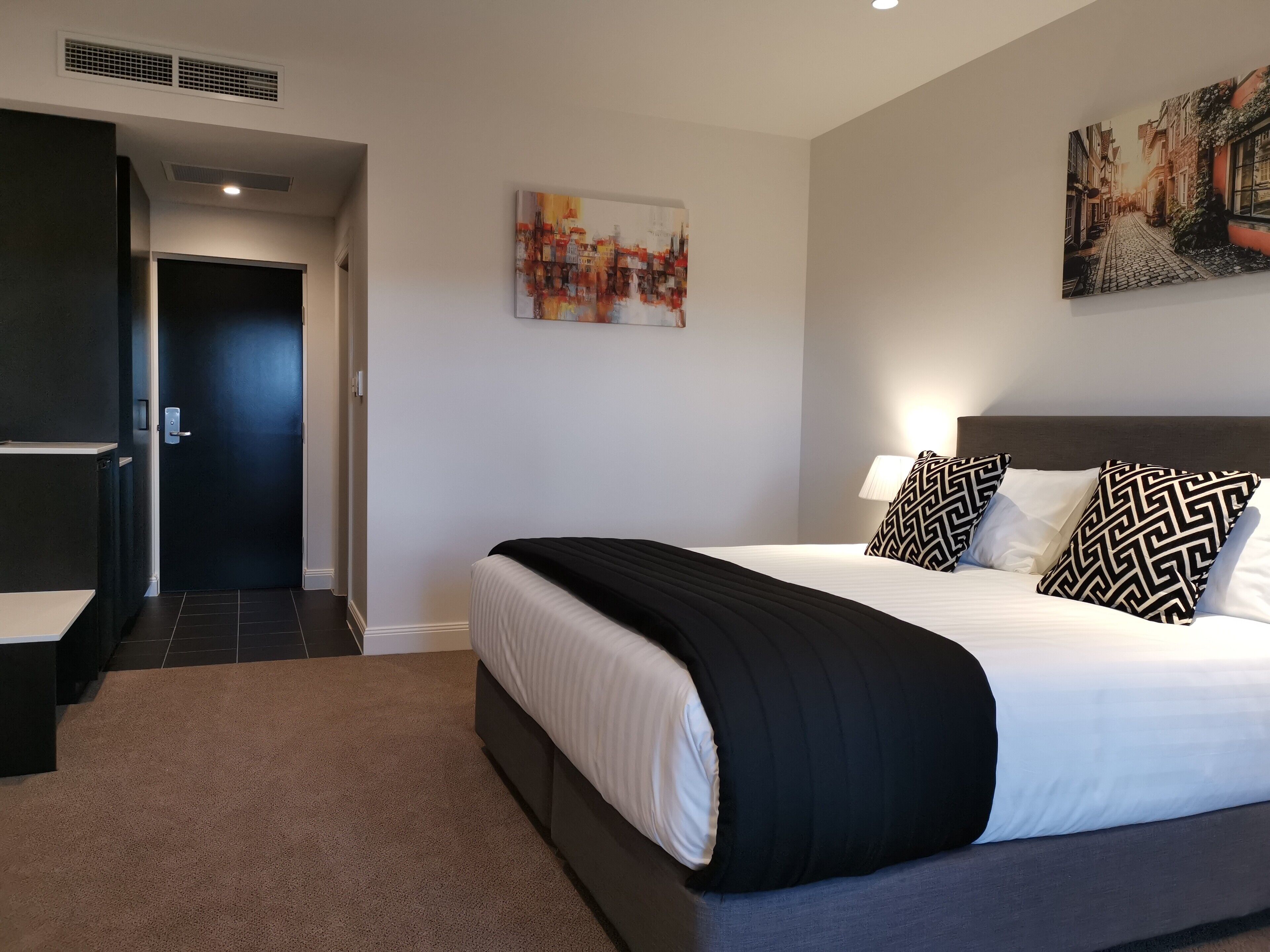 A modern hotel room with a large bed, a nightstand, a painting on the wall, and a door leading to another room.
