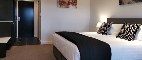 Una habitación de hotel moderna con una cama grande, una mesita de noche, un cuadro en la pared y una puerta que da a otra habitación.