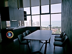 A modern dining area with a large table and chairs, large windows with a city and sea view, and a logo on the wall.
