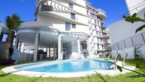 Un bâtiment moderne à plusieurs étages, doté d’une piscine entourée de pelouse et de palmiers.
