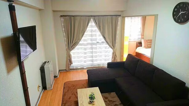 Un espacio habitable compacto con un sofá, una mesita con una planta, un televisor montado en la pared y una ventana con cortinas.