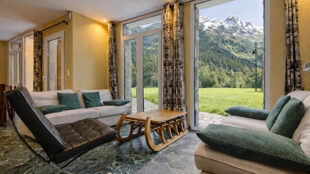 Ein modernes Wohnzimmer mit einer Couch, einem Sessel, einem Couchtisch aus Holz und großen Fenstern mit Blick auf die Berge.