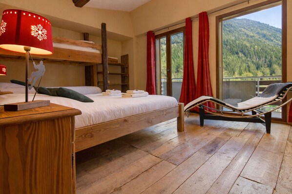 Una habitación con literas, una mesita de noche con lámpara, una silla junto a la ventana y vista a una montaña boscosa.