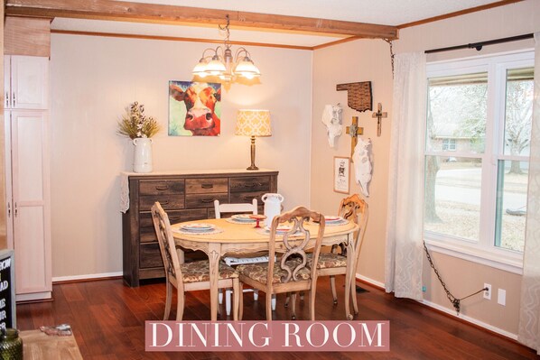 Une salle à manger avec une table en bois, des chaises, un lustre et une fenêtre avec des rideaux.