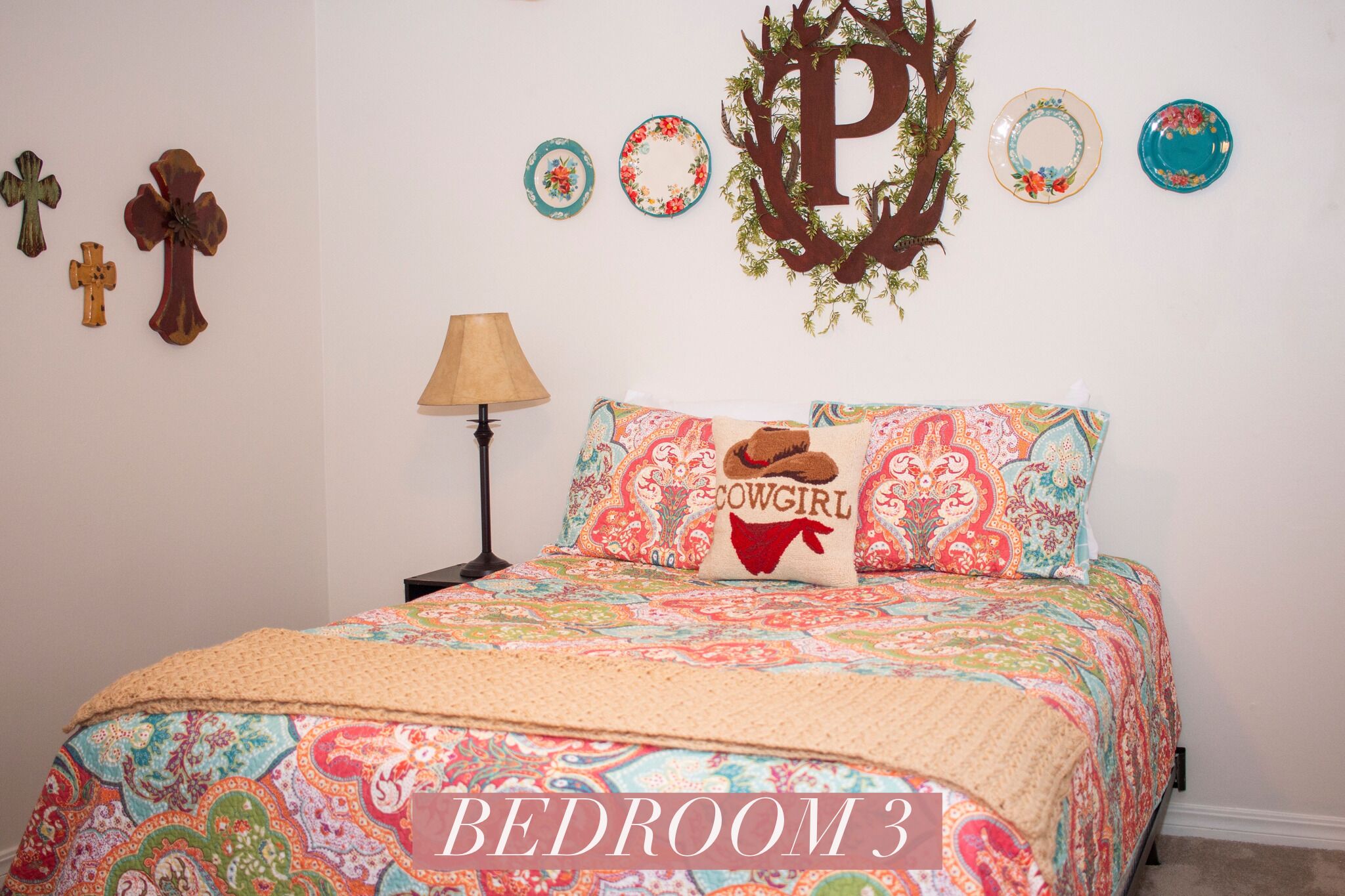 Een slaapkamer met een bed met een geborduurd sprei, een kussen met de tekst 'Cowgirl' en een muur met een grote letter 'P' en decoratieve borden.