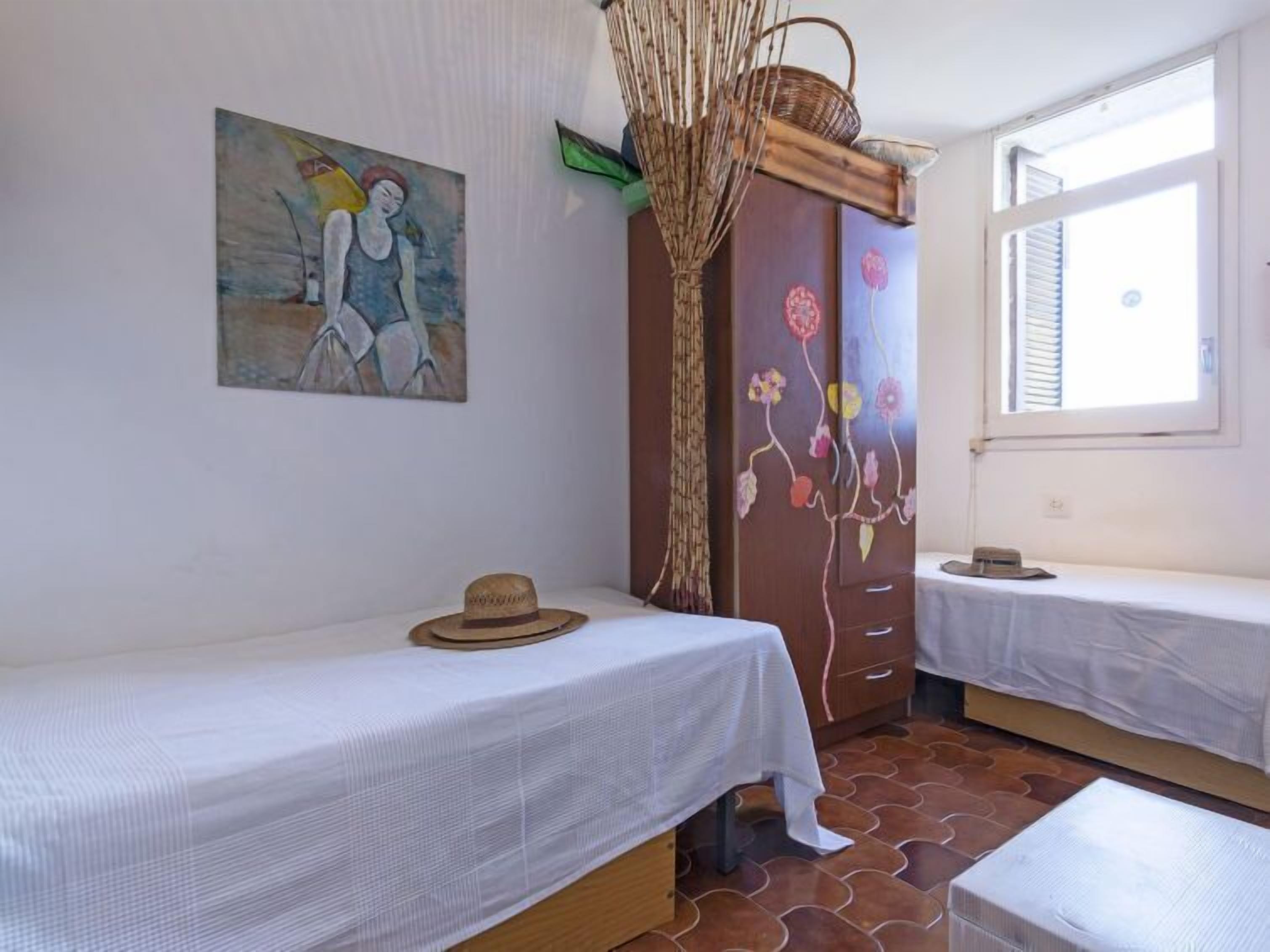 A room with two beds, a painting on the wall, a wooden wardrobe with floral designs, and a window with blinds.