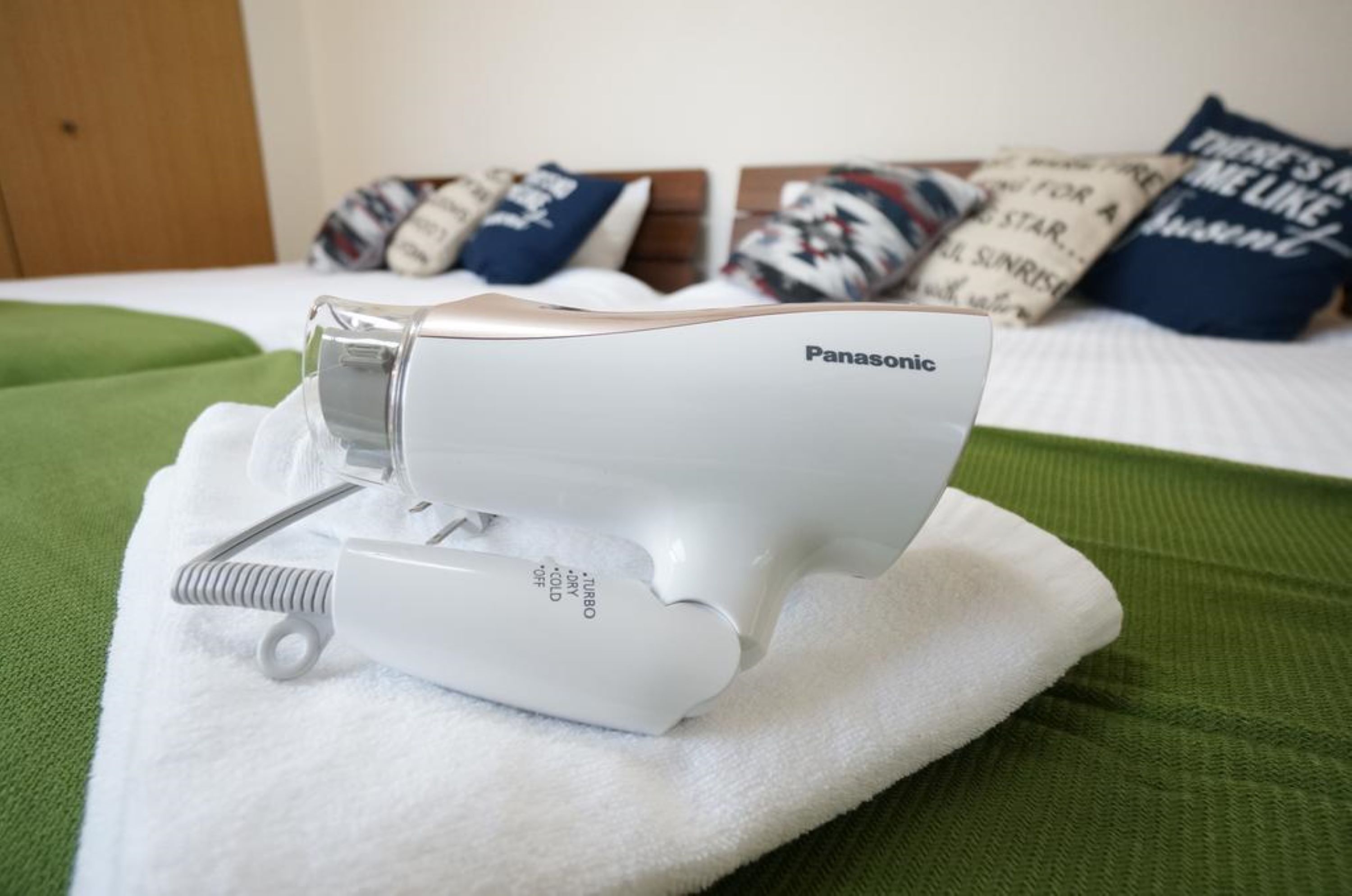 Un secador de pelo Panasonic sobre una toalla blanca en una cama con ropa de cama verde y almohadas estampadas.