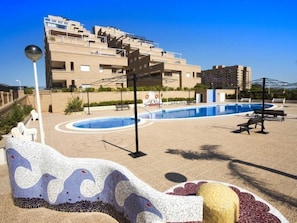 Un espace piscine agrémenté de motifs en mosaïque, un bâtiment doté de nombreux balcons et un ciel d’un bleu limpide.