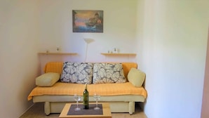A cozy living room with a sofa, two cushions, a small table with a bottle and glasses, and a painting on the wall.